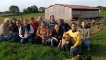 un groupe de personnes accroupies avec des chiens dans un champ avec un bâtiment d'élevage de volailles derrière