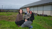 Une femme et un homme accroupis devant un bâtiment d'élevage de volailles avec une poule Coucou de Rennes dan les bras