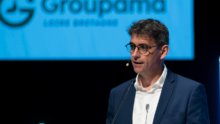 Un homme avec un micro casque à une tribune un écran projette le logo groupama derrière
