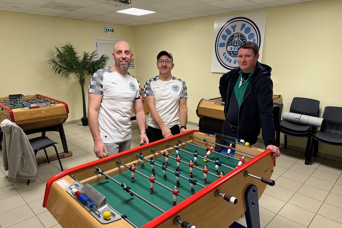 Trois hommes autour d'un baby-foot dans une salle avec plusieurs autres baby-foot - Illustration La passion du baby-foot