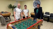 Trois hommes autour d'un baby-foot dans une salle avec plusieurs autres baby-foot