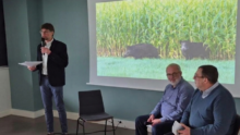 2 hommes assis sur une chaise et un 3ème debout avec un micro devant un grand écran qui projette des sangliers dans un champ de maïs