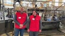 Deux éleveurs habllés d'une veste rouge devant leur manège de traite 28 postes de marque DeLaval