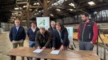 Six hommes dont deux en train de signer des documents sur une table posée dans un bâtiment agricole